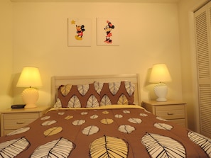 Et soveværelse med sengetæppe med bladmotiver, to vægmonterede lamper og to billeder af Mickey Mouse i rammer.