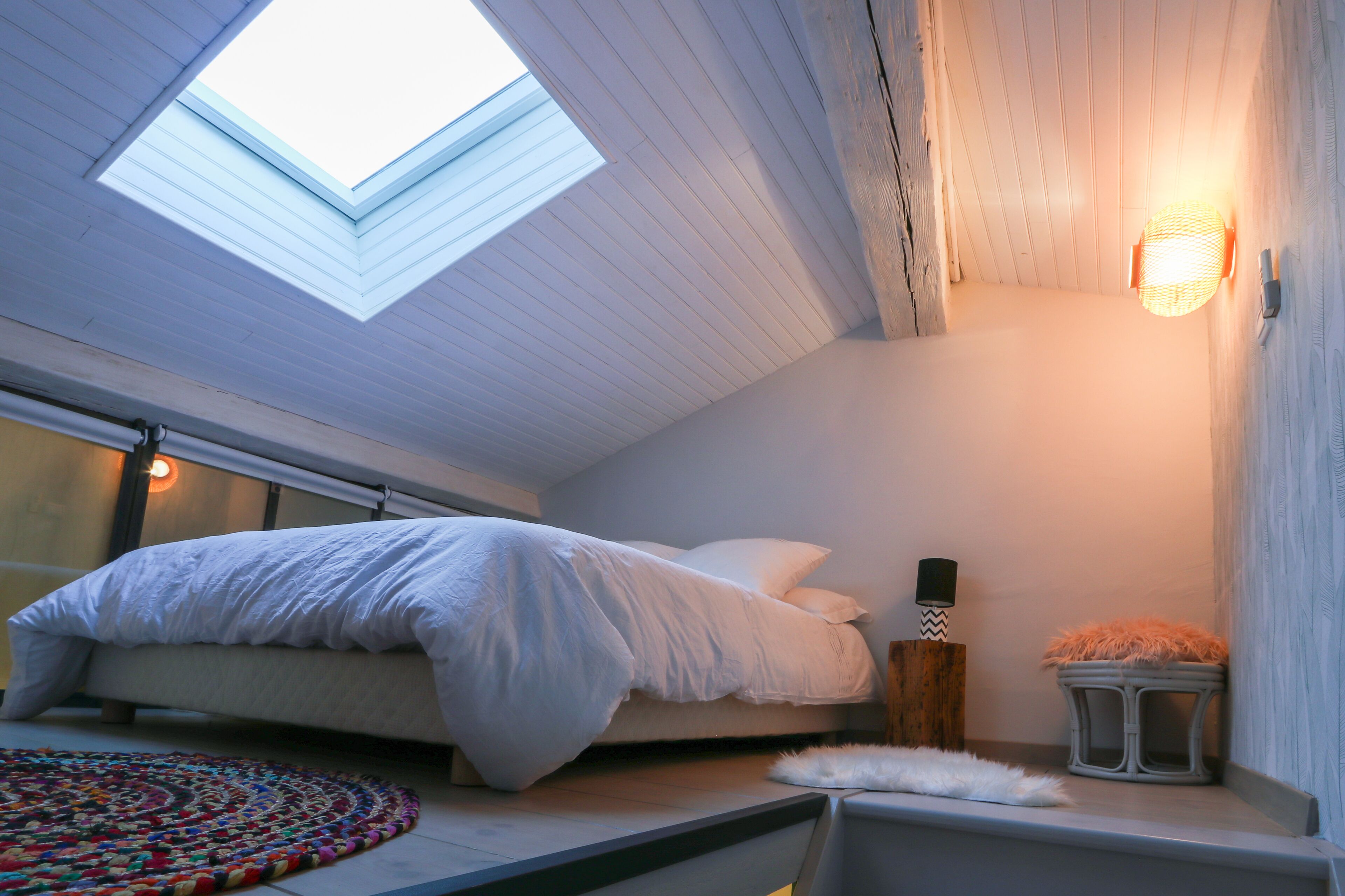 Ein Schlafzimmer mit einer Dachfenster, einem Bett, einem kleinen Hocker und einer Wandleuchte.