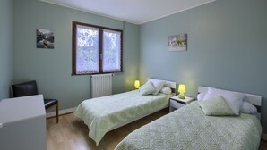 A room with two beds, a chair, a table, and a window with lace curtains.