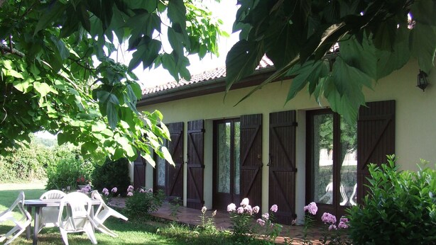 A garden with a table and chairs, a house with shutters, and a pathway.