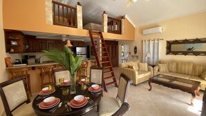 Un coin repas avec une table ronde dressée pour quatre personnes, une cuisine en arrière-plan, un escalier et un salon avec un canapé et une table basse.