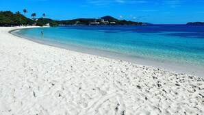 Ein Strand mit klarem türkisblauem Wasser, weißem Sand und einigen Gebäuden im Hintergrund.