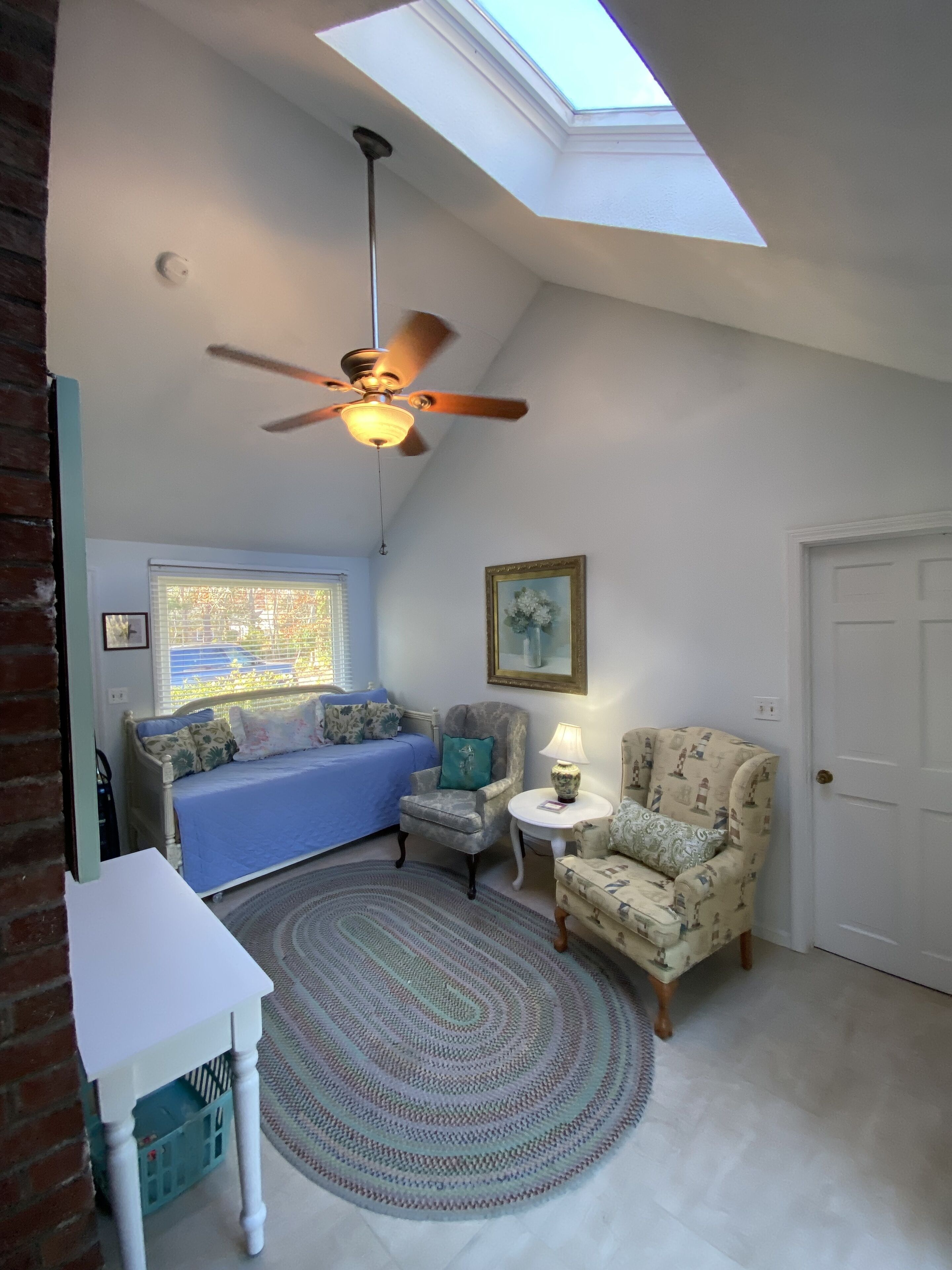 A room with a sloped ceiling, a ceiling fan, a window with blinds, a blue sofa, a patterned armchair, a small table with a lamp, and a white door.