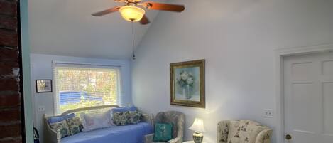 A room with a sloped ceiling, a ceiling fan, a window with blinds, a blue sofa, a patterned armchair, a small table with a lamp, and a white door.