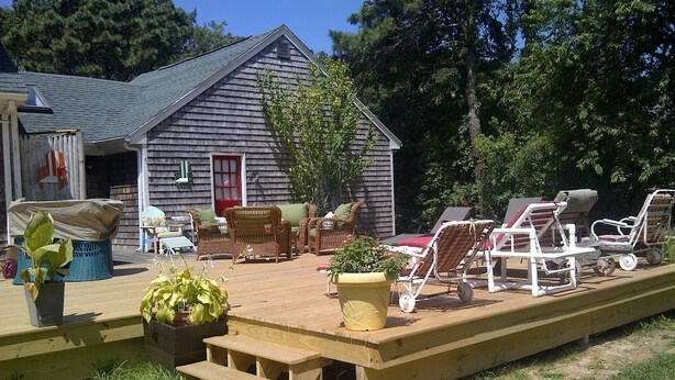 A wooden deck with outdoor furniture, including wicker chairs and lounge chairs, and a small building with a red door in the background. - Quintessential Family Memory Cape Cod Home 1/3 walk to private association beach (East Falmouth)