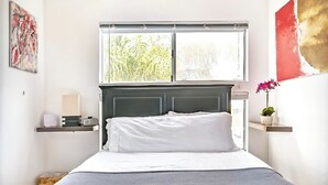 Een slaapkamer met een bed, een raam met zonwering, een nachtkastje met een lamp en een wandplank met een plant.