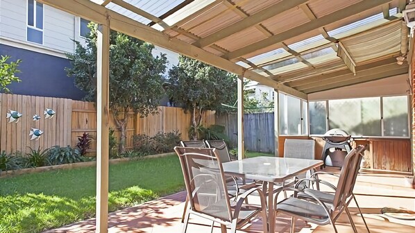 Une terrasse couverte avec une table et des chaises, un barbecue et une clôture en bois en arrière-plan.