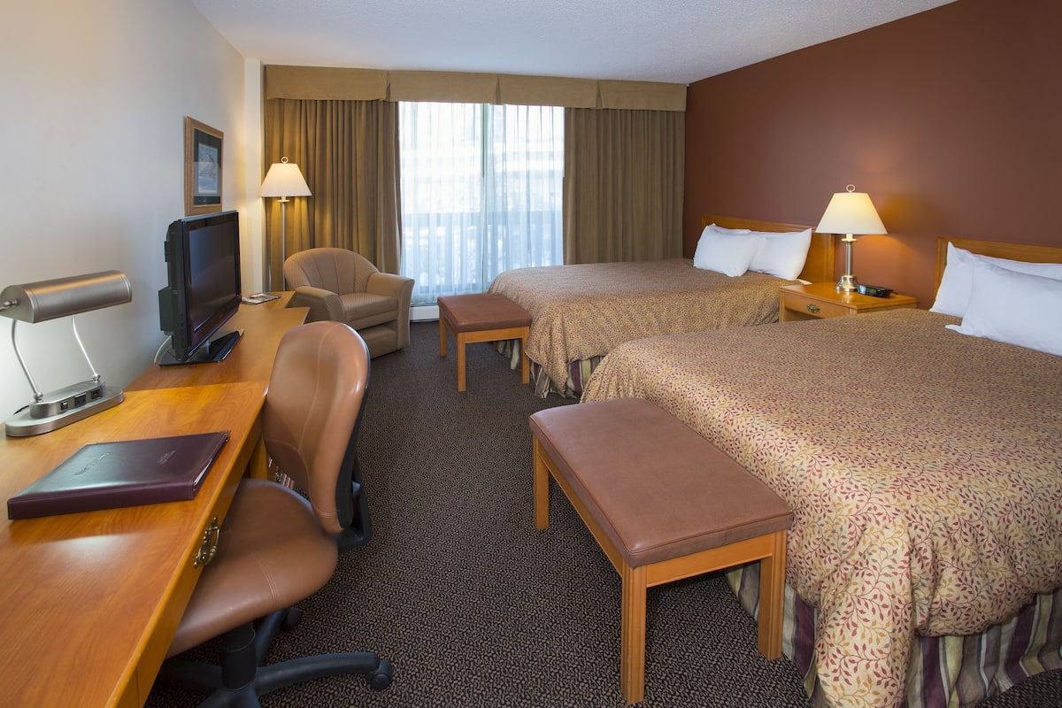 A hotel room with a desk, two beds, a chair, a TV, and a window with curtains.