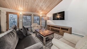 A modern living room with a wooden ceiling, a large flat-screen TV, a wooden coffee table, and a beige sofa.