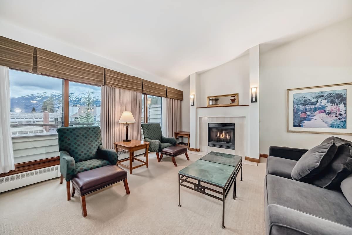 A hotel room with a fireplace, a sofa, a chair, a coffee table, and a view of a snowy mountain.