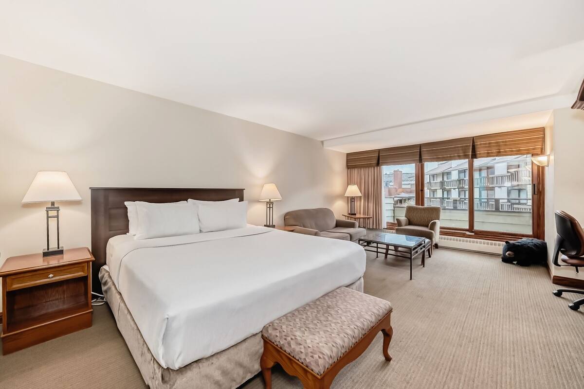 A hotel room with a large bed, two bedside lamps, a sofa, a small table, and a view of buildings through the window.