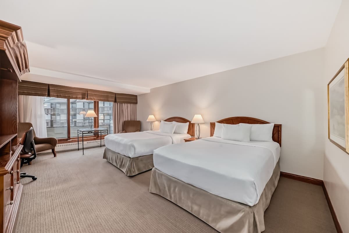 A hotel room with two beds, a desk, a chair, and a window with a view of buildings.