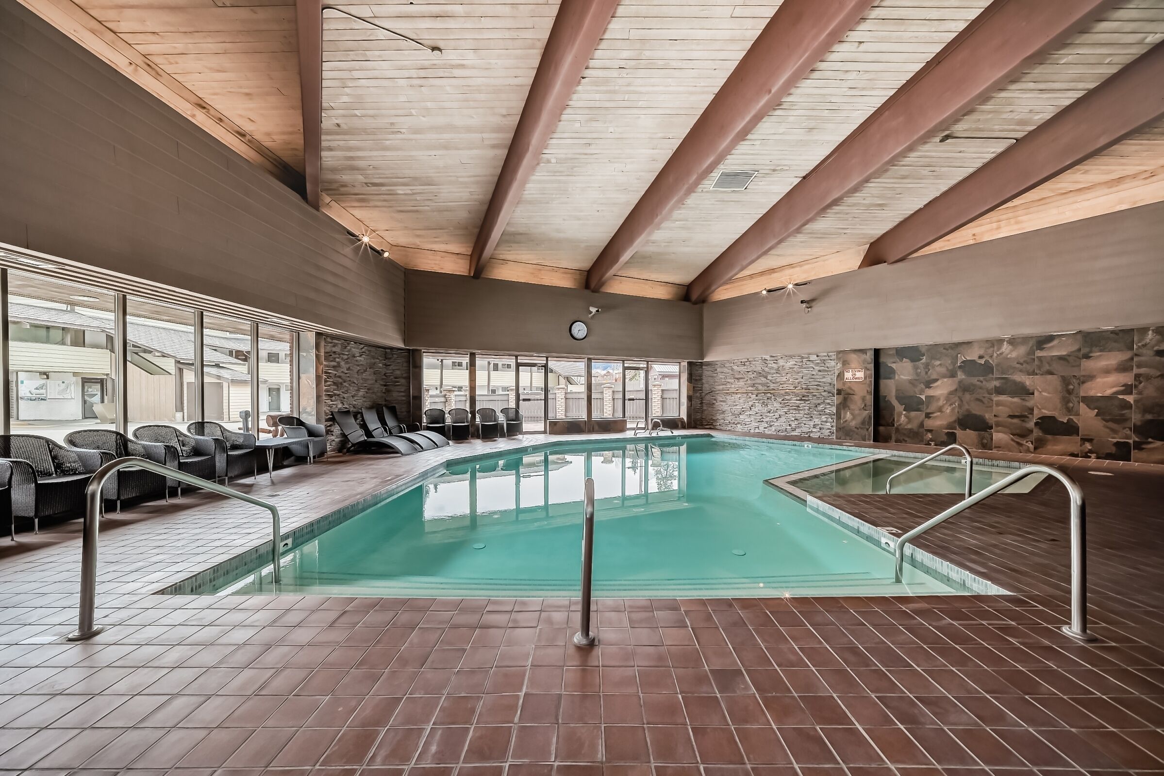 An indoor swimming pool with tiled flooring, metal railings, and a stone wall backdrop.