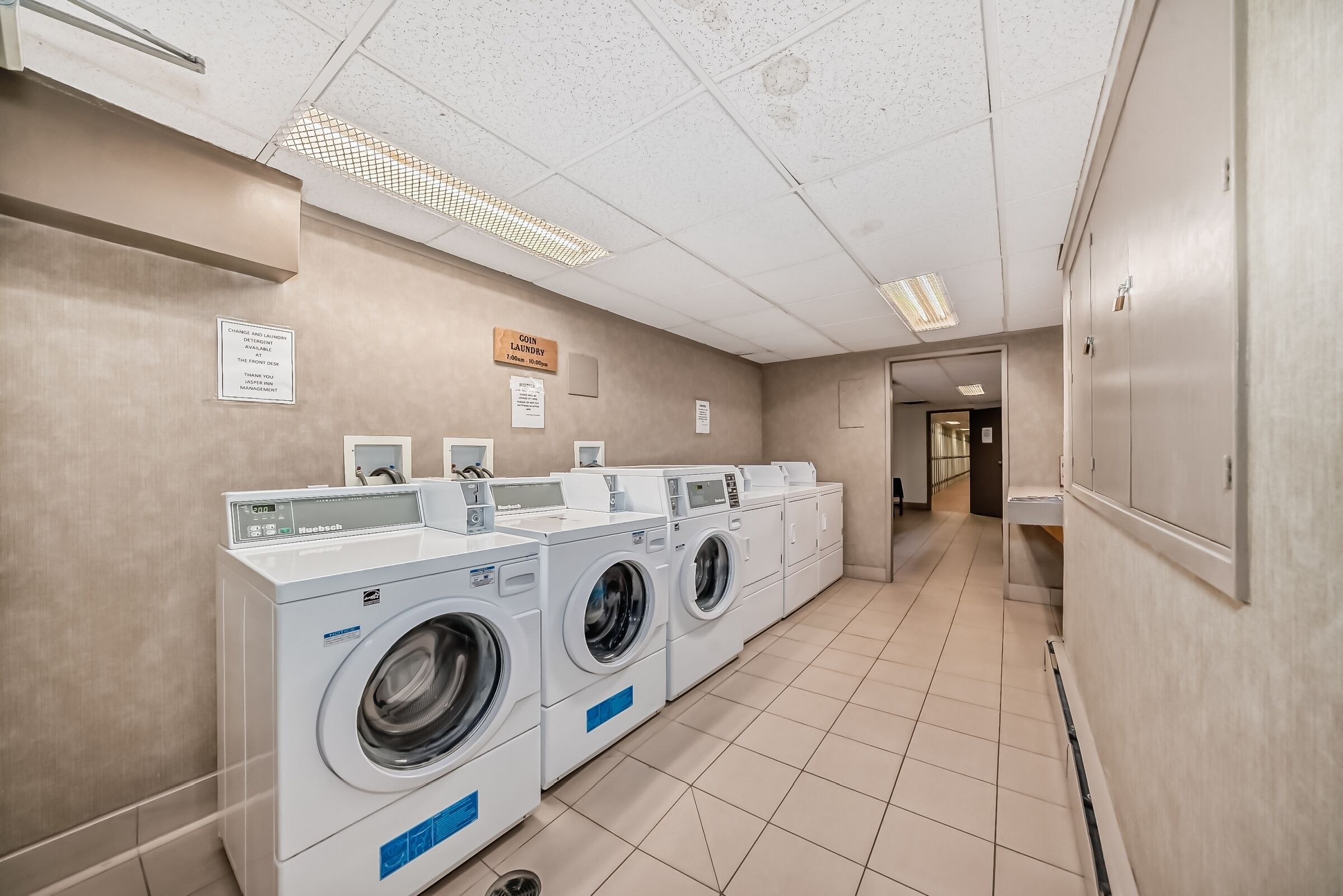 A laundry room with multiple washing machines and dryers, a sign indicating the location, and a tiled floor.