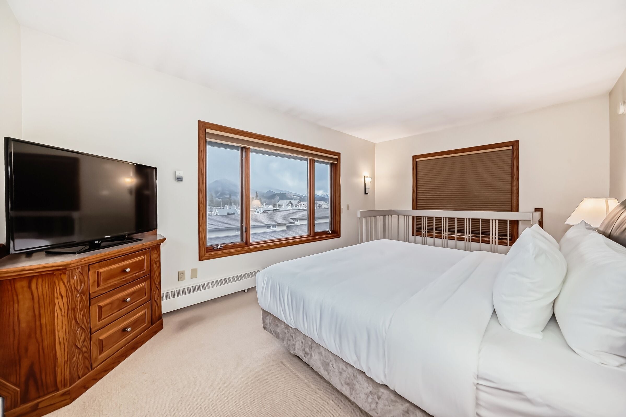 Un dormitorio con una cama grande, un televisor sobre un aparador de madera, una ventana con vistas a edificios y un radiador.