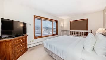 Un dormitorio con una cama grande, un televisor sobre un aparador de madera, una ventana con vistas a edificios y un radiador.
