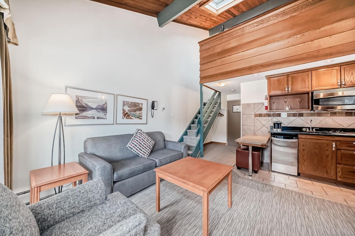 A modern living room with a grey sofa, a wooden coffee table, and a staircase leading to a kitchen with wooden cabinets and stainless steel appliances.
