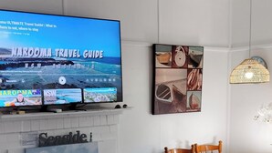 Un salon avec un téléviseur diffusant un guide de voyage, un coin repas avec une table et des chaises, et un ventilateur de plafond.