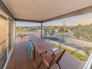 A balcony with a wooden table and chairs, a glass railing, and a view of a road and buildings.