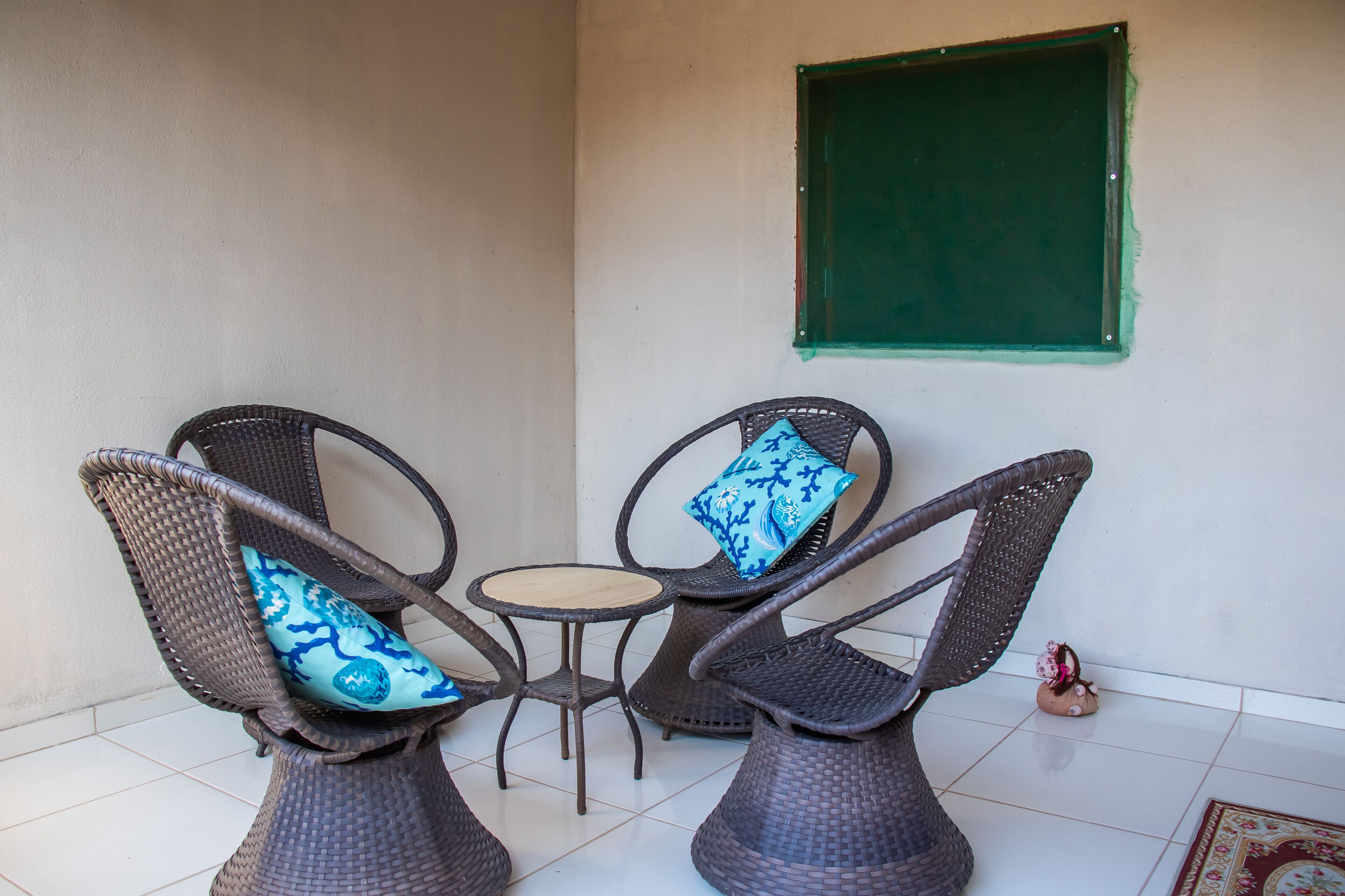 Een zithoek met twee rieten stoelen en een klein rond tafeltje, een raam met groene omlijsting, en een betegelde vloer.