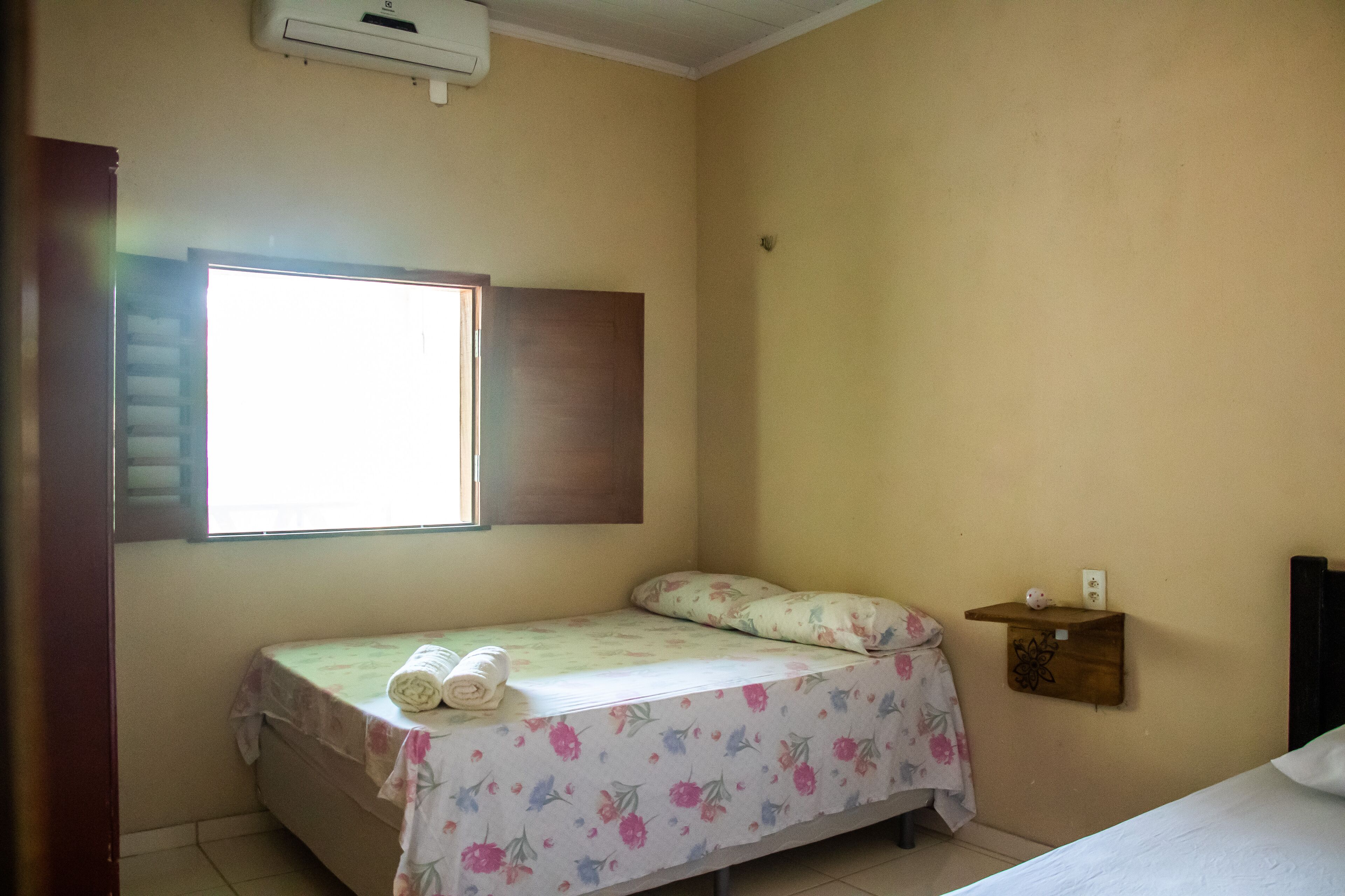 A small, single-bed room with a window, a wooden shelf with a decorative item, and a wall-mounted air conditioner.