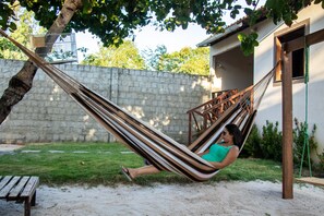 A hammock tied between two trees in a backyard with a stone wall, a wooden bench, and a small building with a window. - CASA MANU - PREÁ JERI (Cruz)