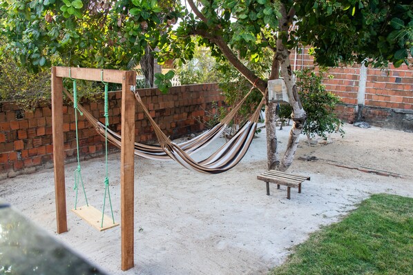 A hammock swing set with a wooden bench nearby, set against a brick wall and surrounded by trees. - CASA MANU - PREÁ JERI (Cruz)