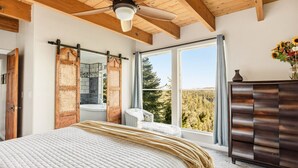 Une chambre avec un grand lit, un plafond en bois, une fenêtre donnant sur les arbres, une commode et un ventilateur de plafond.