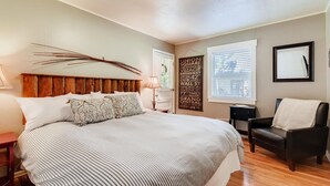 Une chambre à coucher avec un grand lit, une tête de lit en bois, une table de chevet, une chaise, une fenêtre avec des stores et un mur sur lequel est affichée une plaque indiquant les noms des rues.