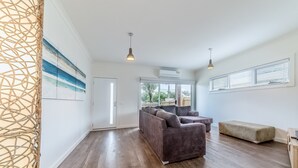 Amplia sala de estar con un sofá modular grande, un banco y piso de madera. Se ve una puerta que da a otra habitación, una ventana grande con persianas y un ventilador de techo.