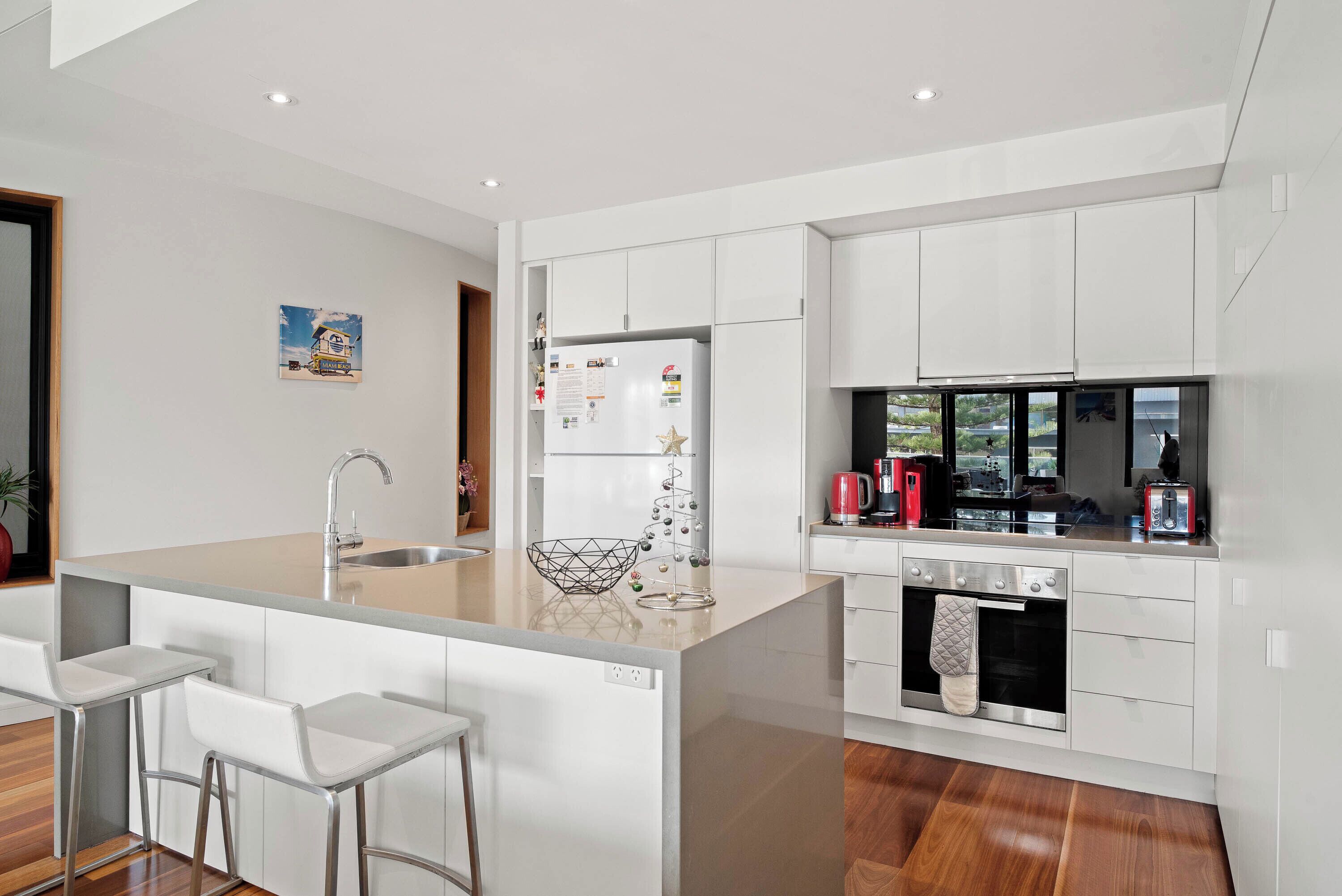 A modern kitchen with white cabinetry, a central island with bar stools, a built-in oven, and a window overlooking a parking area.