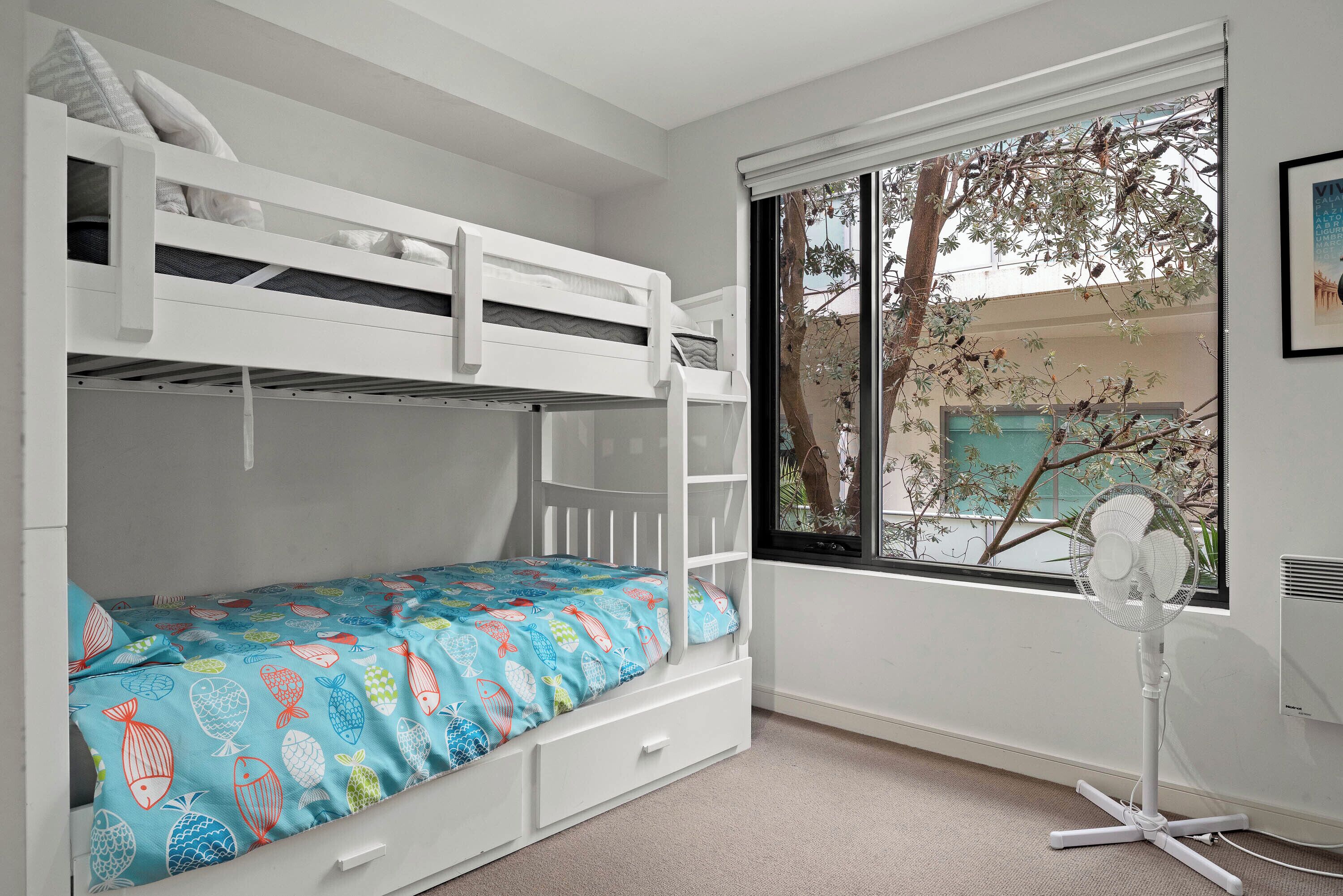 A bunk bed with a blue fish-patterned bedspread, a white fan, and a window with a view of trees.