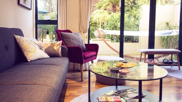Een woonkamer met een bank, een rode fauteuil, een glazen salontafel en uitzicht op een tuin door grote ramen.