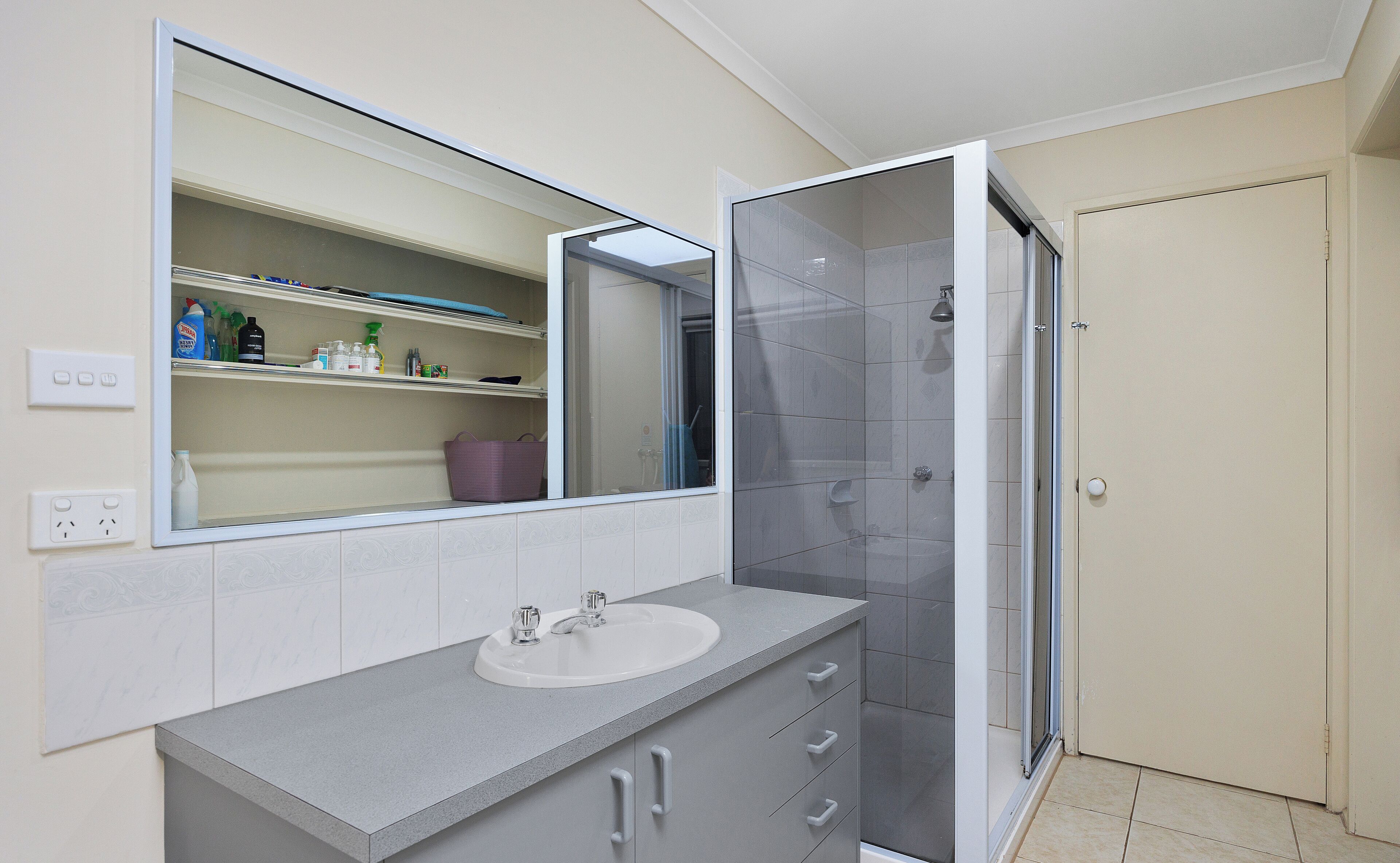 A bathroom with a large mirror, a sink, a shower, and a storage shelf with toiletries.