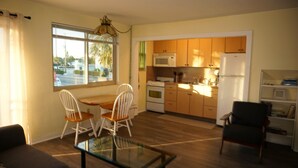 A compact kitchen with wooden cabinets, a white refrigerator, and a microwave. There's a dining table with two chairs and a glass-top coffee table. - 1BR beachside condo ~ pool, beach, restaurants!  Balcony with gulf views. FN 806 (St Pete Beach)