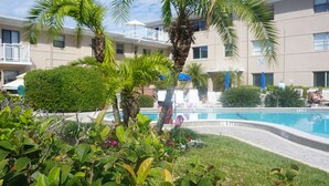A pool area with lounge chairs, palm trees, and a building with balconies in the background. - 1BR beachside condo ~ pool, beach, restaurants!  Balcony with gulf views. FN 806 (St Pete Beach)
