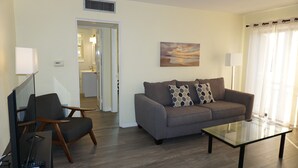 A living room with a sofa, armchair, and glass coffee table. A painting hangs on the wall, and a kitchen is visible through an open door. - 1BR beachside condo ~ pool, beach, restaurants!  Balcony with gulf views. FN 806 (St Pete Beach)