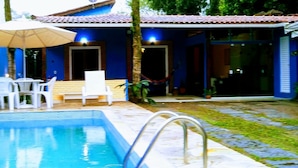 A swimming pool with a white chair and an umbrella, a covered patio area, and a small garden with a hammock.