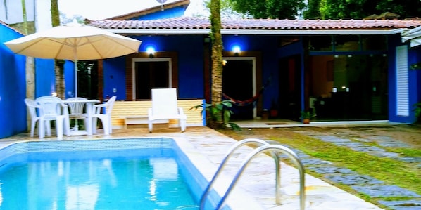 A swimming pool with a white chair and an umbrella, a covered patio area, and a small garden with a hammock.