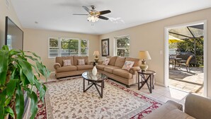 A living room with a brown sofa set, a glass coffee table, and a ceiling fan. There are two lamps on side tables, and a large window with a view of the outdoors. - Perfect for big family reunions, sport teams! (Fort Myers)