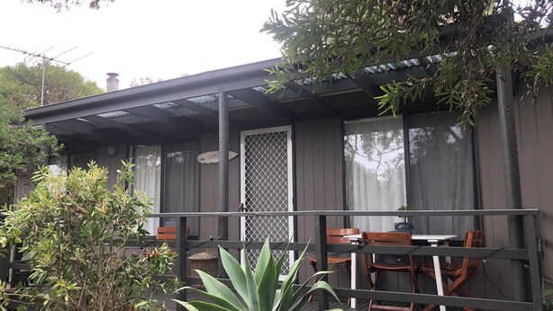 Uma área de estar coberta ao ar livre, com cadeiras e mesa de madeira, cercada por plantas e um portão de metal.