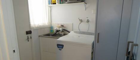 A compact kitchen with a sink, a washing machine, and a shelf with toiletries.
