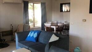 Uma sala de estar com um sofá cinza escuro, um banquinho branco e uma mesa de jantar com cadeiras.