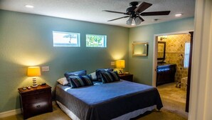 A bedroom with a bed, two bedside lamps, a ceiling fan, and a bathroom visible through an open door.