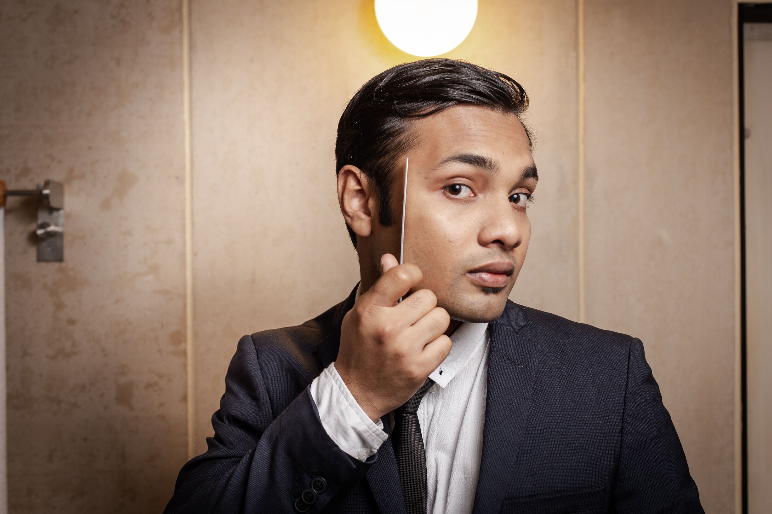 A man in a dark suit adjusting his tie in a room with a light fixture on the wall.