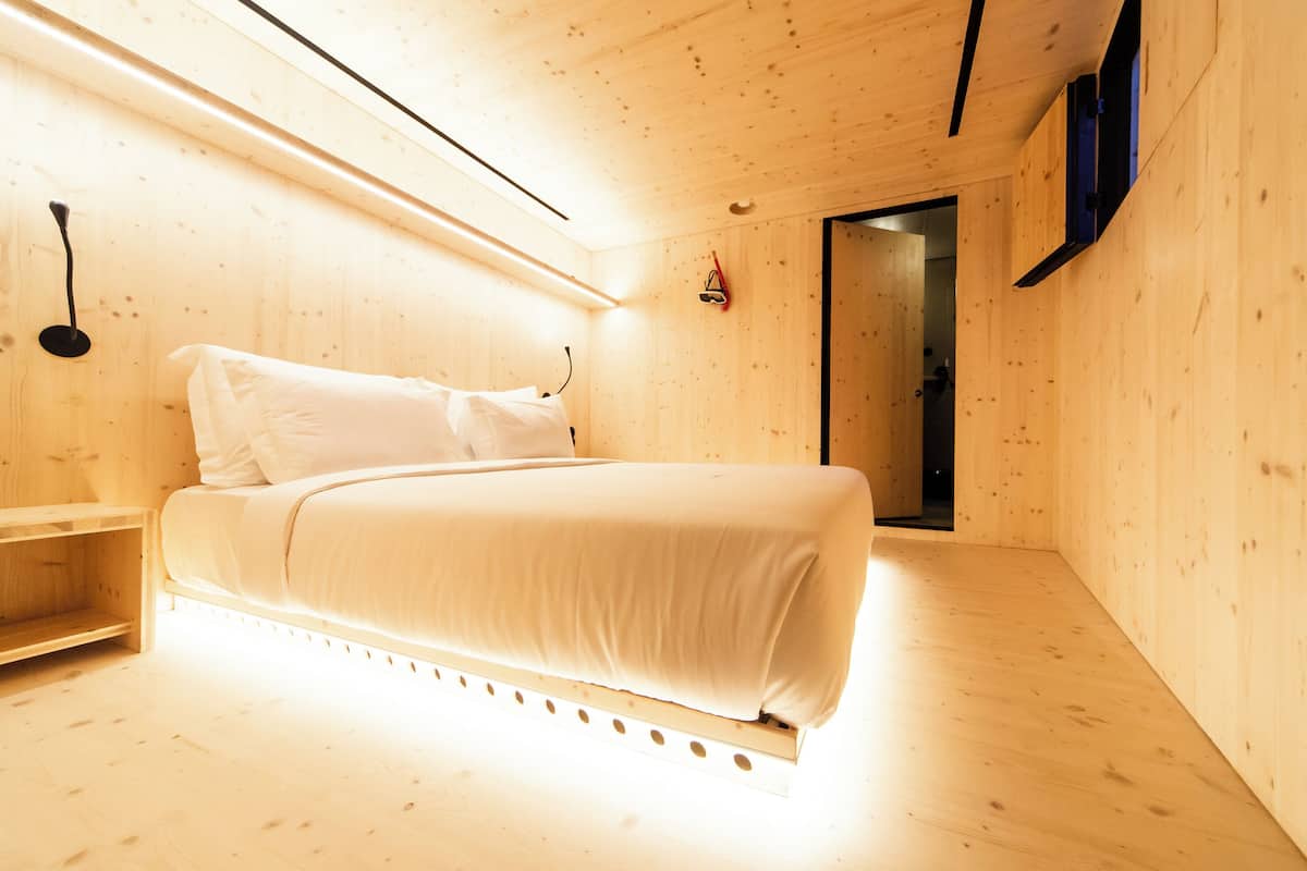 A minimalist wooden room with a bed, a bedside table, and a wall-mounted light fixture.