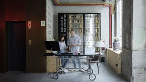 Un mostrador de recepción con una computadora portátil, un hombre y una mujer de pie cerca, y una vitrina con objetos en su interior.