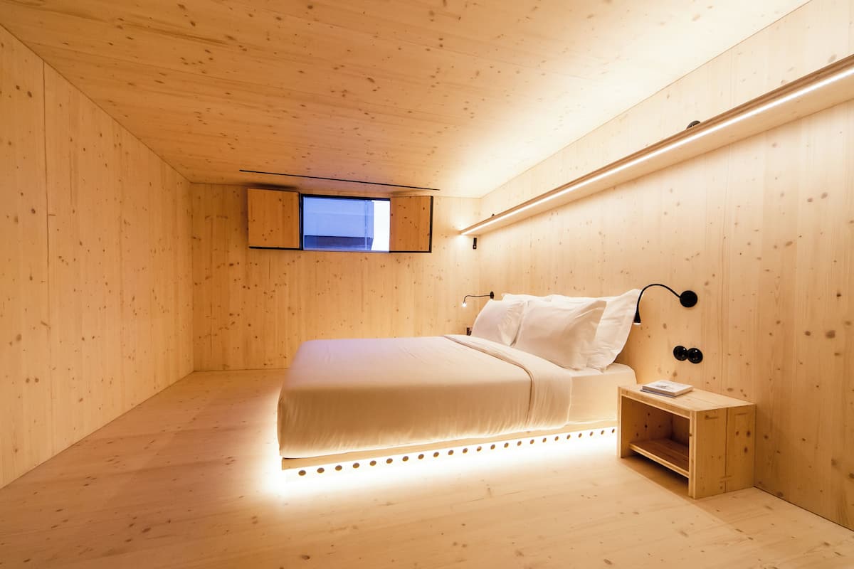 A minimalist wooden room with a bed, a small bedside table, and a window.