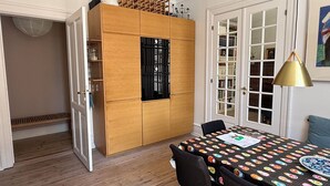 Un'area pranzo con un tavolo ricoperto da una tovaglia fantasia, sedie, un mobiletto in legno con ripiani e una lampada a sospensione.