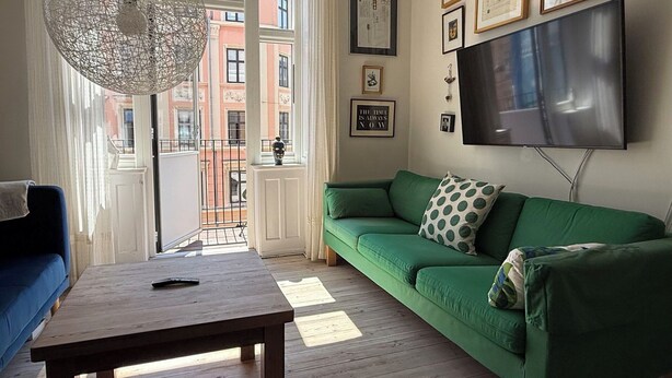 Un soggiorno con un divano verde, un tavolino in legno, una televisione e un'ampia finestra con vista sugli edifici.